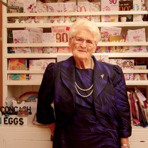 Elderly white woman photographed from the hips up against a background of shelving displaying cards