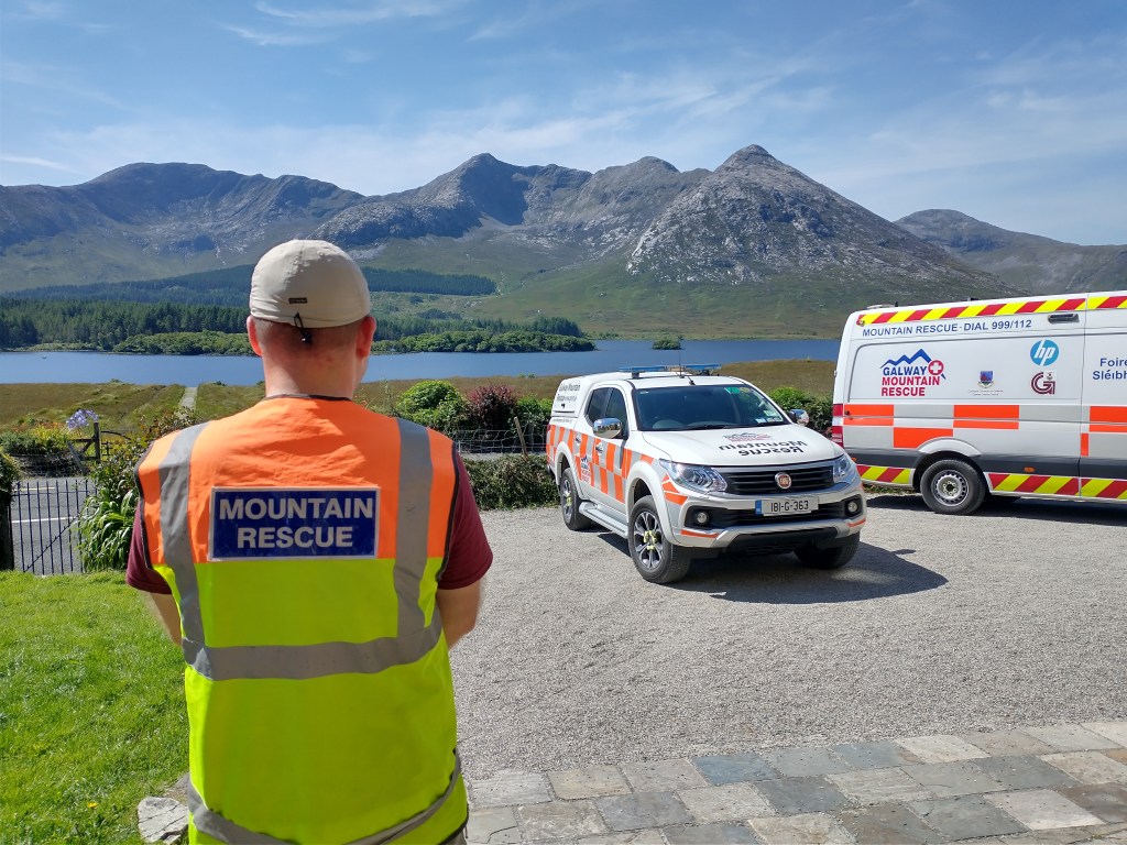 Galway Mountain Rescue Team member facing mountains, a lake, and two rescue vehicles.