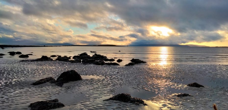 Image of Ballyloughane beach with blue and gold sunset sky and low tide exposing rocks in the sand