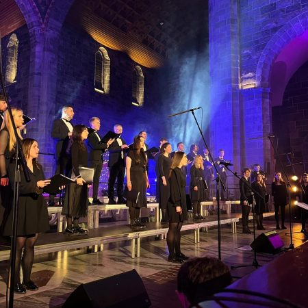 Claddagh Choral members standing on a blue-lit stage holding song books and singing