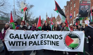 Members of the Galway branch of the IPSC are standing behind a white banner that reads 'Galway Stands with Palestine'. The banner also has the circular logo of the IPSC which is made up of the Palestinian flag in the middle and the words 'Ireland-Palestine Solidarity Campaign' around it. They are marching on the streets of Dublin in a National Demonstration for Palestine. People behind the banner are holding Palestinian flags.