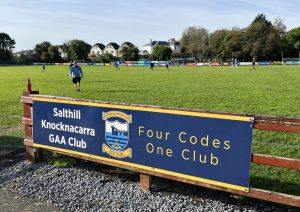 Image shows Salthill Knocknacarra GAA Prairie pitch. In the foreground of the image we see blue signage attached to the wooden fence around the pitch. The signage has the words Salthill Knocknacarra GAA Club and the clubs logo. On the right it states 'Four Codes One Club". There are some people playing sports casually on the pitch