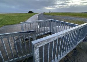 In for foreground is a kissing gate, which is a U-shaped steel barrier with a hinged swinging gate in the middle. The swinging gate means only one side is open at any time. In the background is a walking and cycle coastal path through a green park