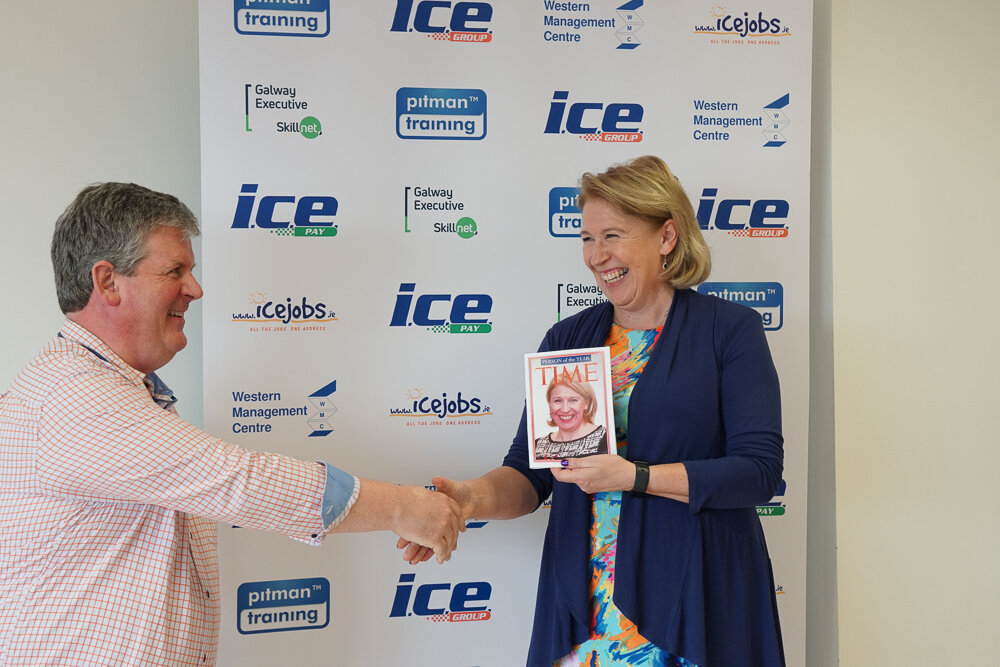 Felim McDonnell, a man wearing a pink shirt and Margaret Cox wearing a navy suit jacket of ICE Group are shaking hands in front of a promotional banner. The banner shows the logos of local businesses and business organisations. Margaret is holding a mock issue of Time magazine with a picture of her on the cover. Both are shown from the waist up and are laughing.