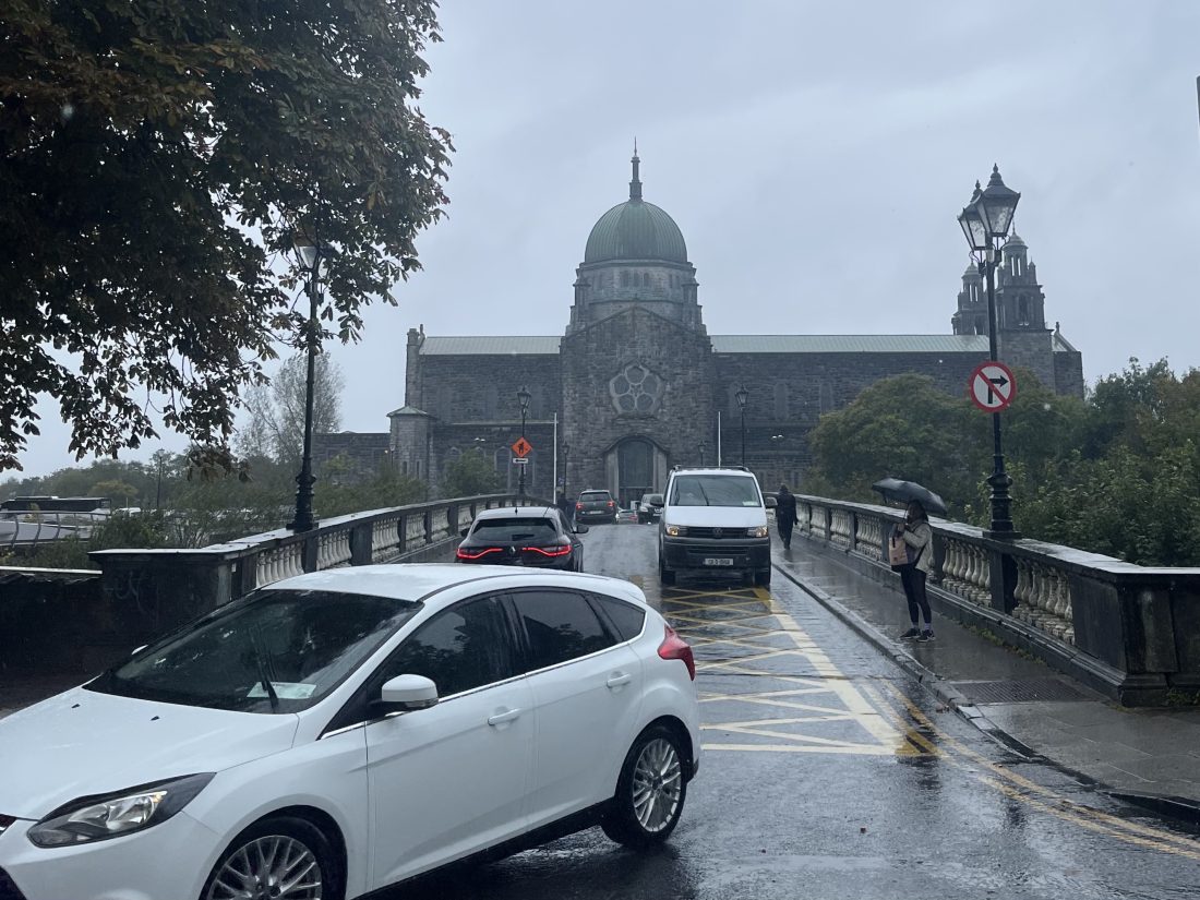 Cars and pedestrians on a bridge with Galway cathedral in the background. There's a white car in the foreground turning off the bridge