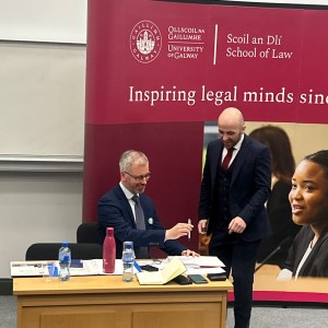 Minister Roderic O'Gorman at an event in the University of Galway. He's standing beside a table with a maroon University banner behind him. There's another man Dr Brian Tobin sitting at the desk. Both are wearing suits
