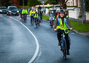 A group of adults and children are cycling to school in a line on a road. In the foreground, is a Marshall leading the cycle. She's wearing a hi-vis and a helmet.