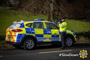 A Garda (Irish policeman) standing beside a Garda car on a main road checking for speeding cars. The Garda car is coloured with blue and luminous yellow squares
